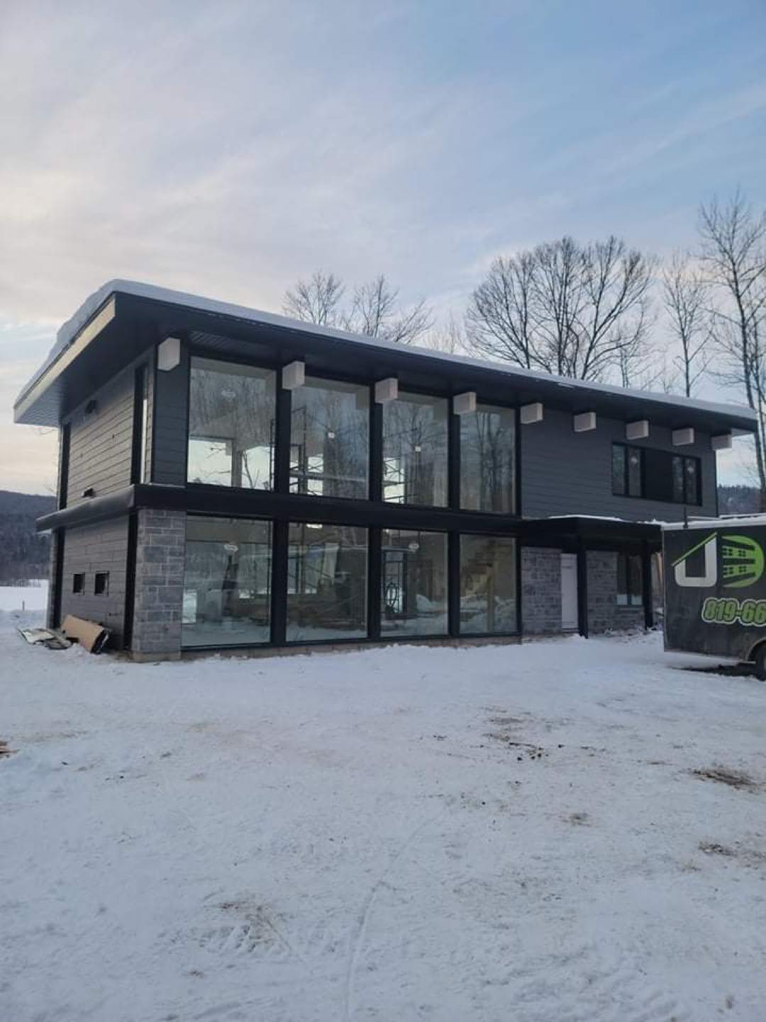 Maison moderne à deux étages avec de grandes fenêtres, un revêtement extérieur gris et des éléments en pierre, située dans un paysage enneigé.