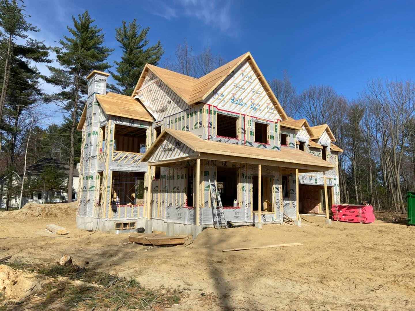 Maison à deux étages en construction, à ossature bois avec bardage en contreplaqué, toit apparent, fenêtres et porche, par une journée ensoleillée.