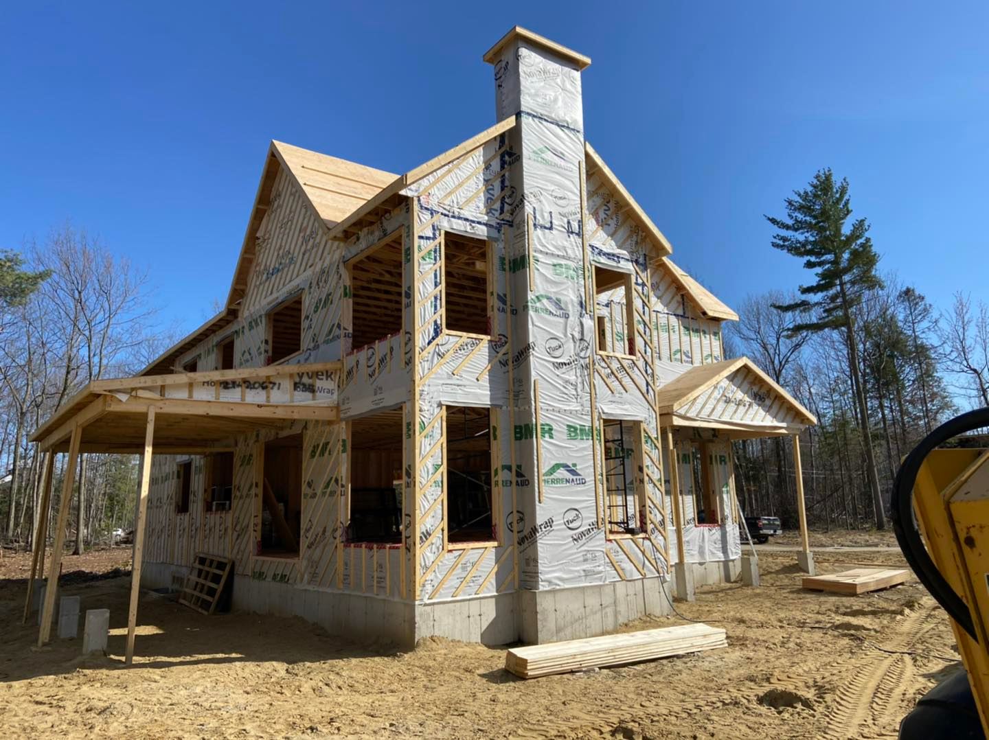 Maison à deux étages en construction, avec charpente apparente et grande cheminée. Porche visible sur fond de ciel bleu.