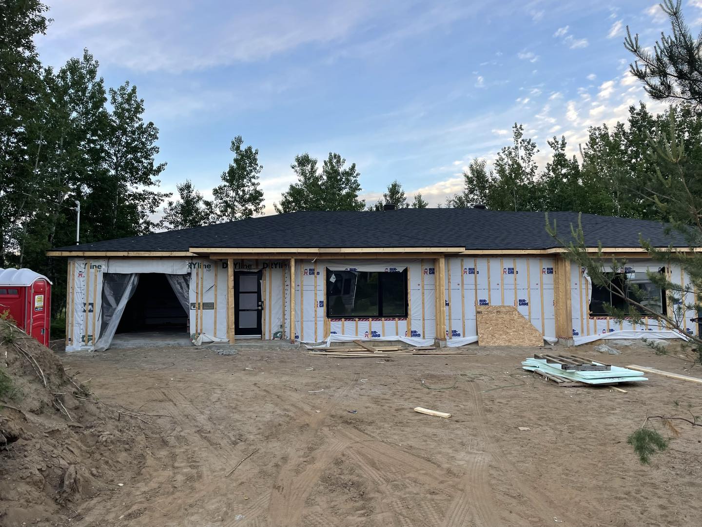 Maison neuve en construction, toit sombre et bardage apparent. Garage ouvert, ciel bleu.