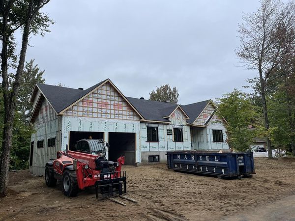 Une maison en construction avec un chariot élévateur rouge et une benne à ordures bleue à l'extérieur, par temps nuageux.