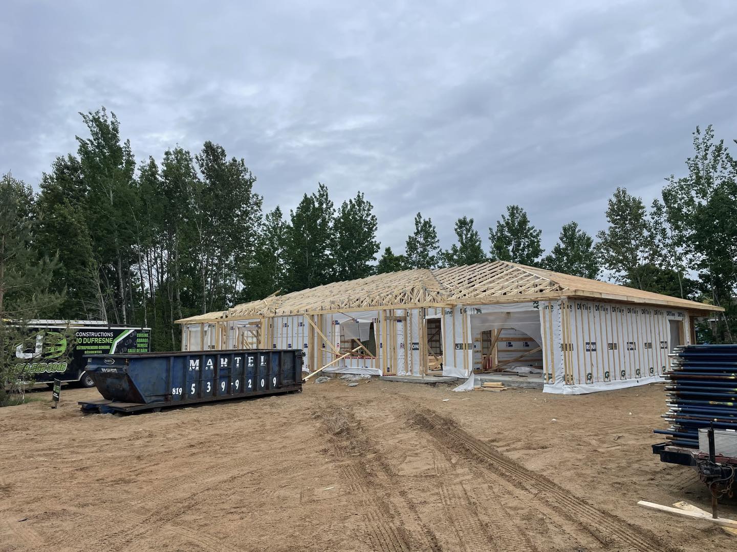 Construction d'une maison de plain-pied. On aperçoit la charpente en bois, le contreplaqué et une partie de la toiture. Une benne à ordures bleue est posée sur le sol.