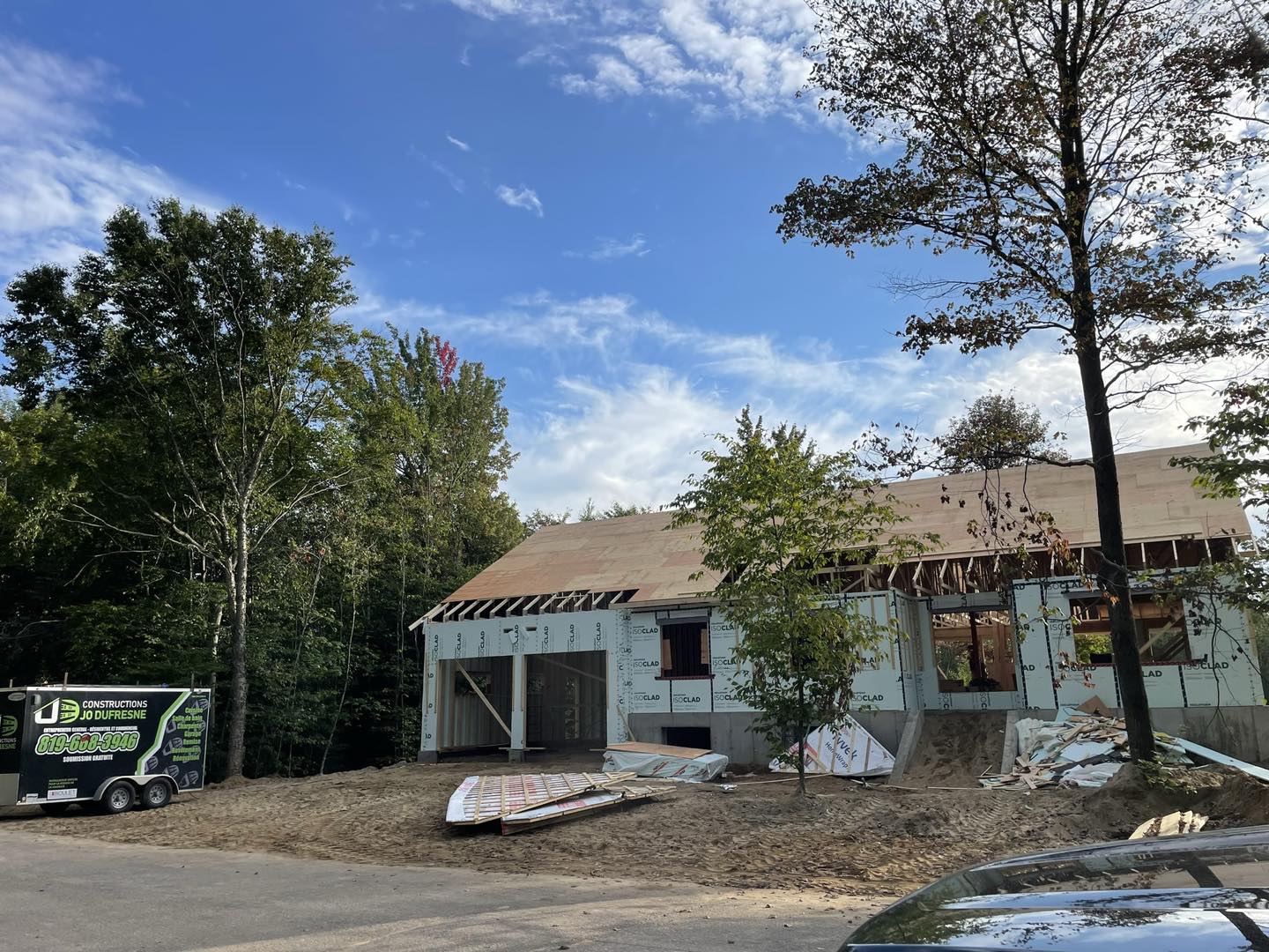 Maison en construction à charpente apparente, entourée d'arbres sous un ciel bleu.