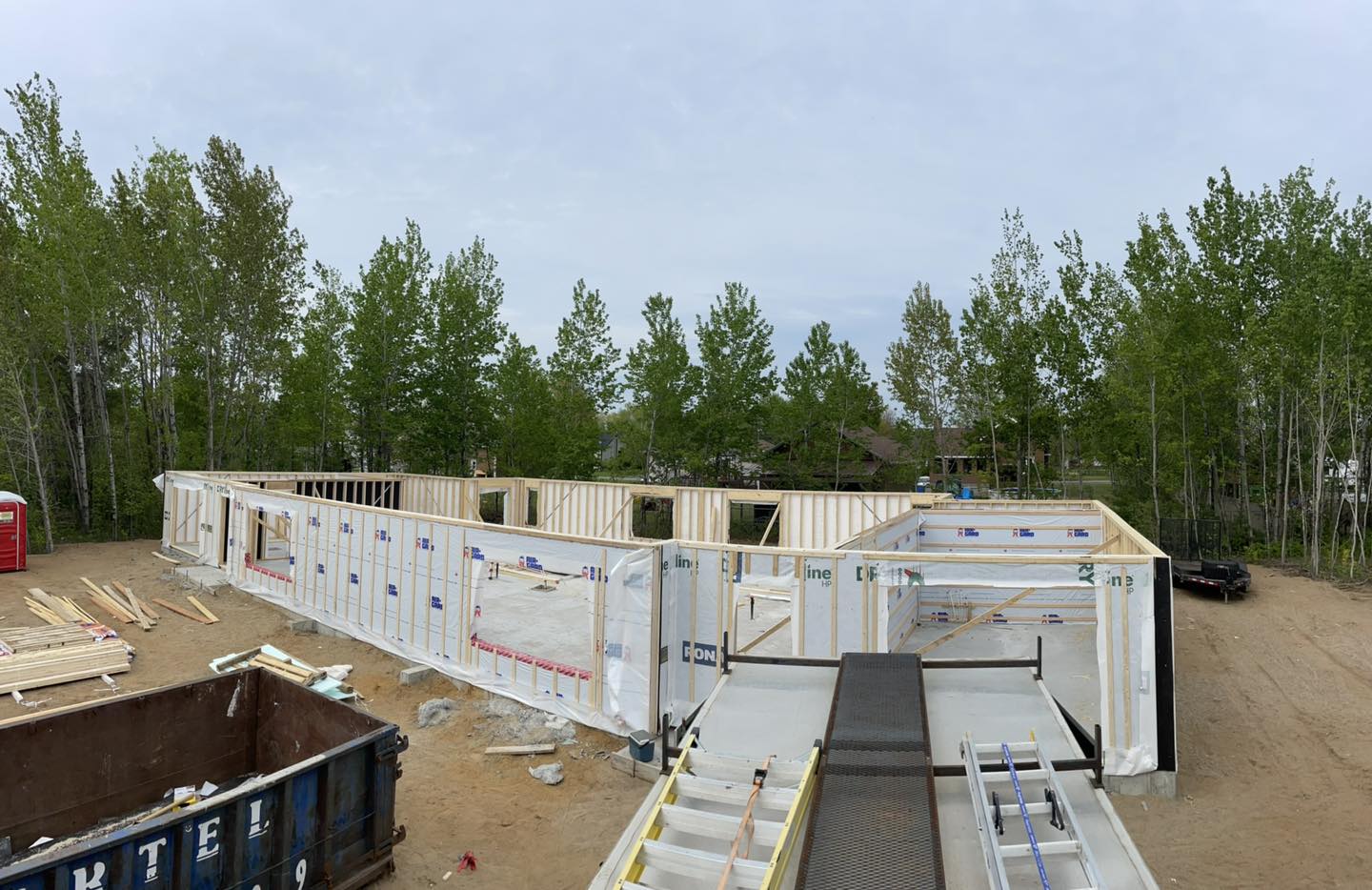 Chantier de construction avec murs à ossature bois ; panneaux d'isolation extérieure installés ; entouré d'arbres et d'un ciel nuageux.