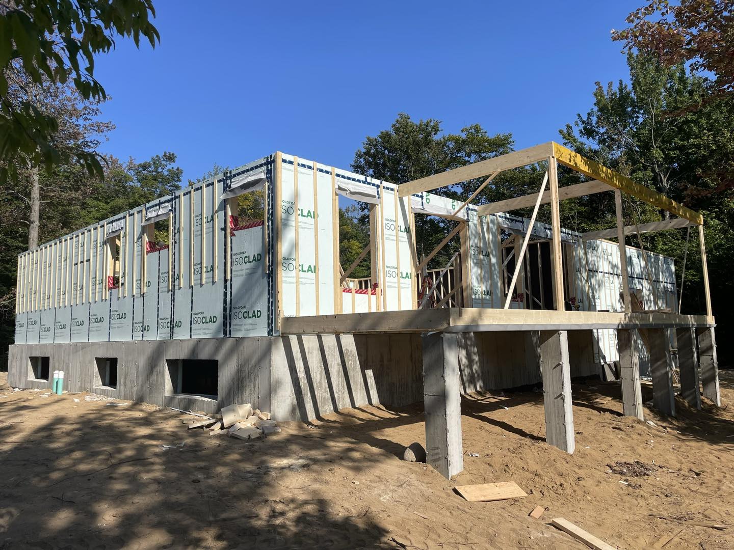 Chantier de construction : ossature bois d'une maison sur piliers en béton, sous un ciel bleu, entourée d'arbres.