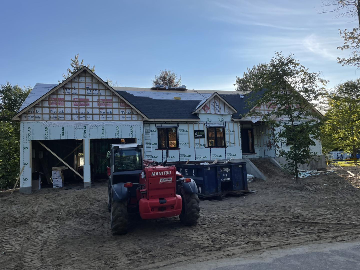 Une maison en construction avec un chariot élévateur rouge devant ; des toilettes portables bleues se trouvent près de l'entrée.