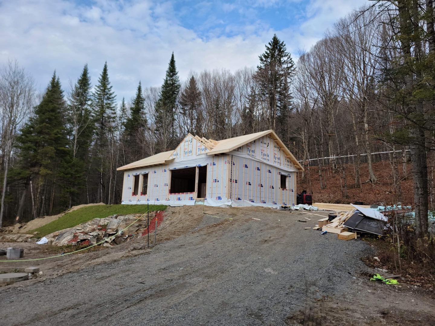 Maison en construction sur une allée de gravier, entourée d'arbres et sous un ciel partiellement nuageux.