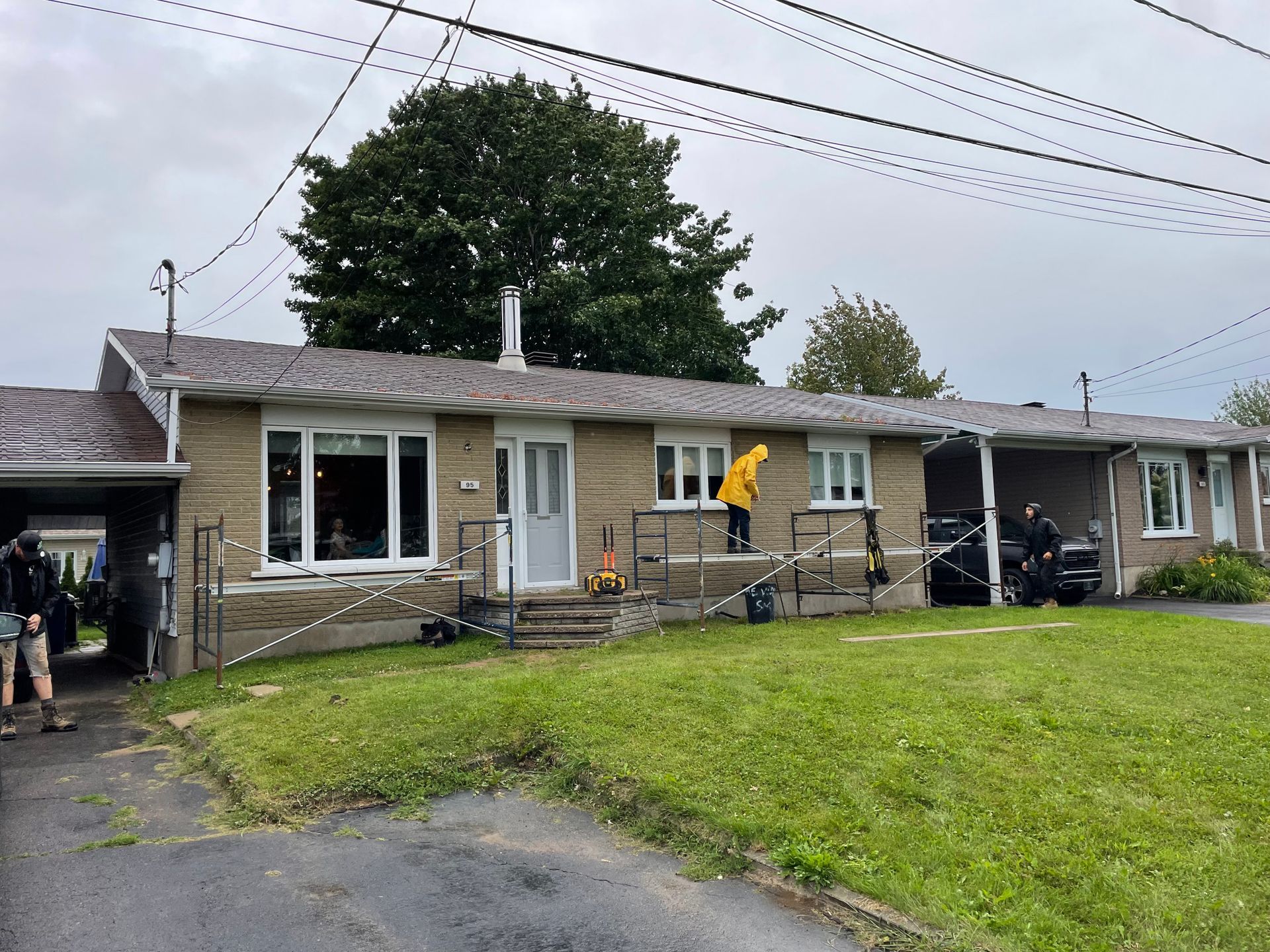 Une maison de plain-pied en briques avec un abri voiture couvert. Une personne vêtue d'un imperméable jaune se tient sur le perron.