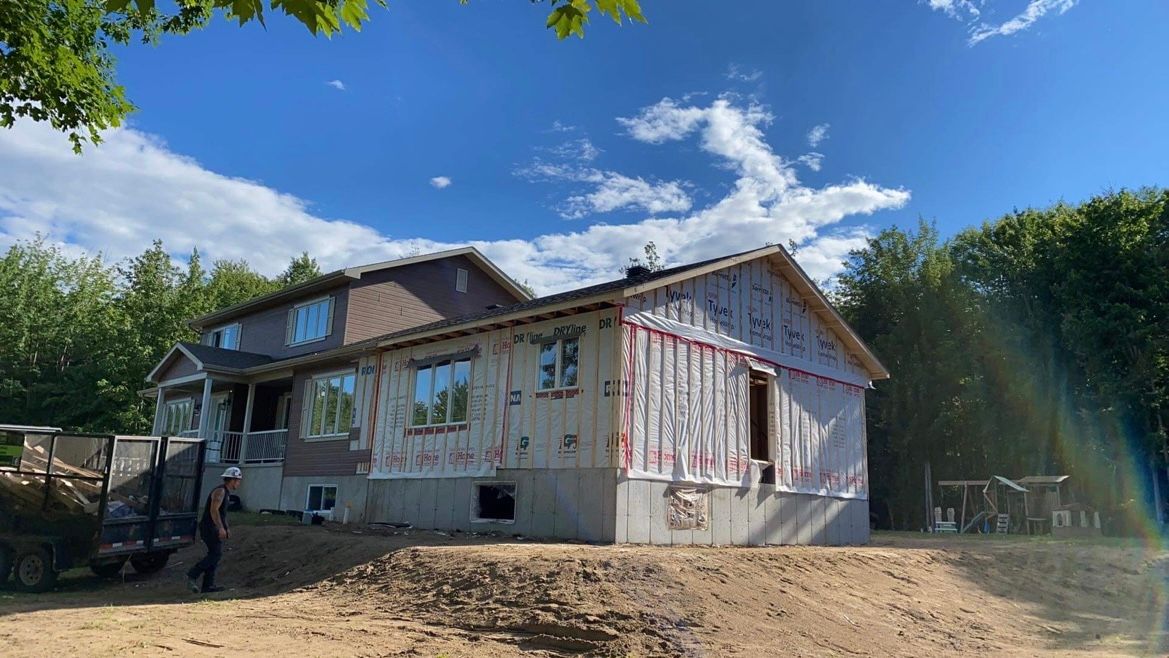 Maison en construction par une journée ensoleillée. Présence d'une personne et d'une remorque.