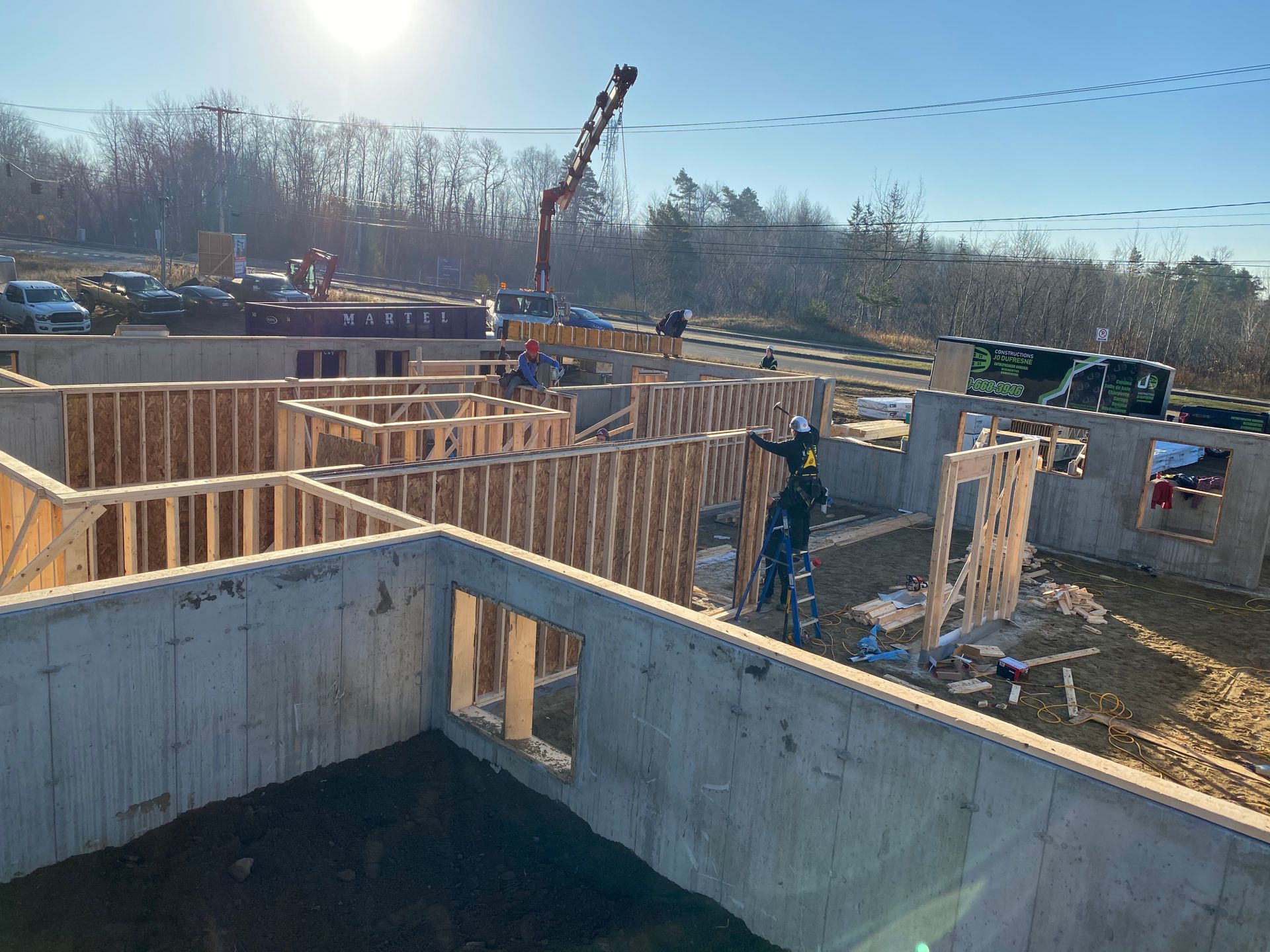 Chantier de construction avec des ouvriers en train de charpenter un bâtiment. Fondations en béton, murs en bois, grue en arrière-plan.