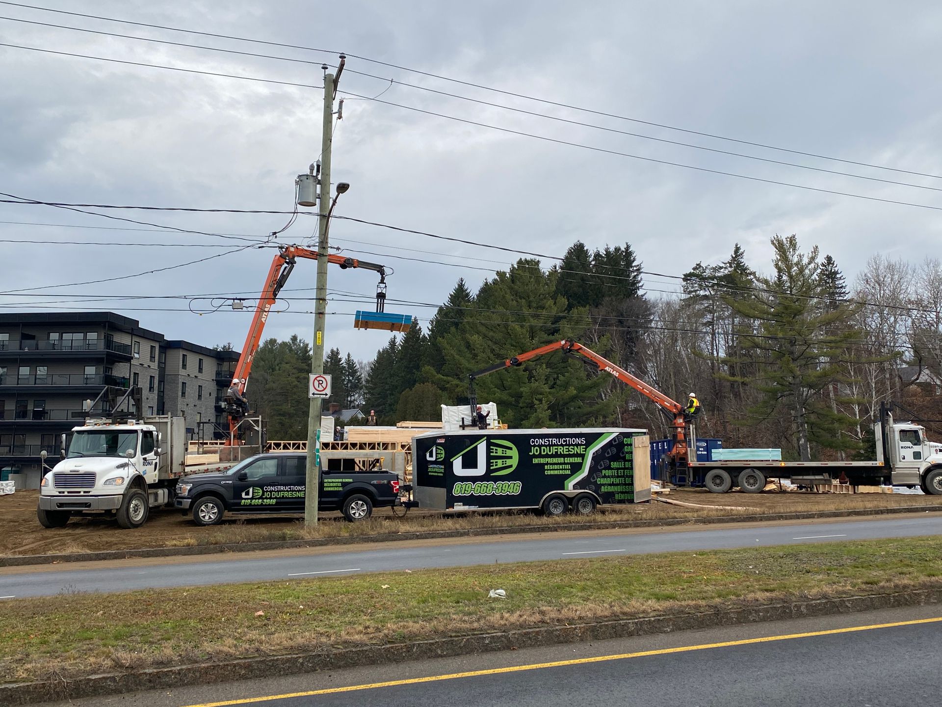 Chantier de construction avec des grues soulevant des matériaux. Des camions, une remorque et un bâtiment en construction sont visibles sous un ciel nuageux.