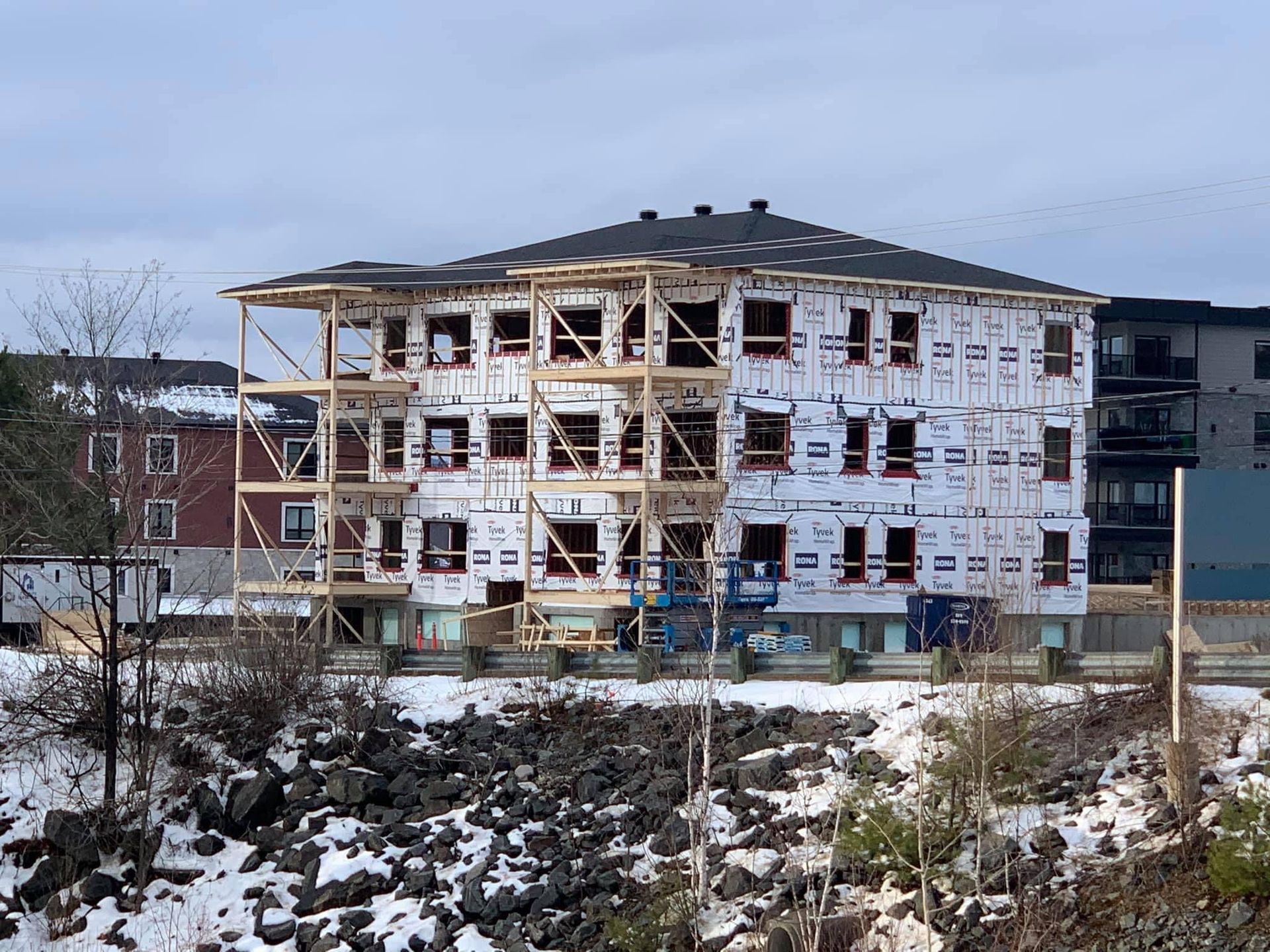 Bâtiment à plusieurs étages en construction, vue extérieure ; charpente en bois, revêtement blanc, neige.