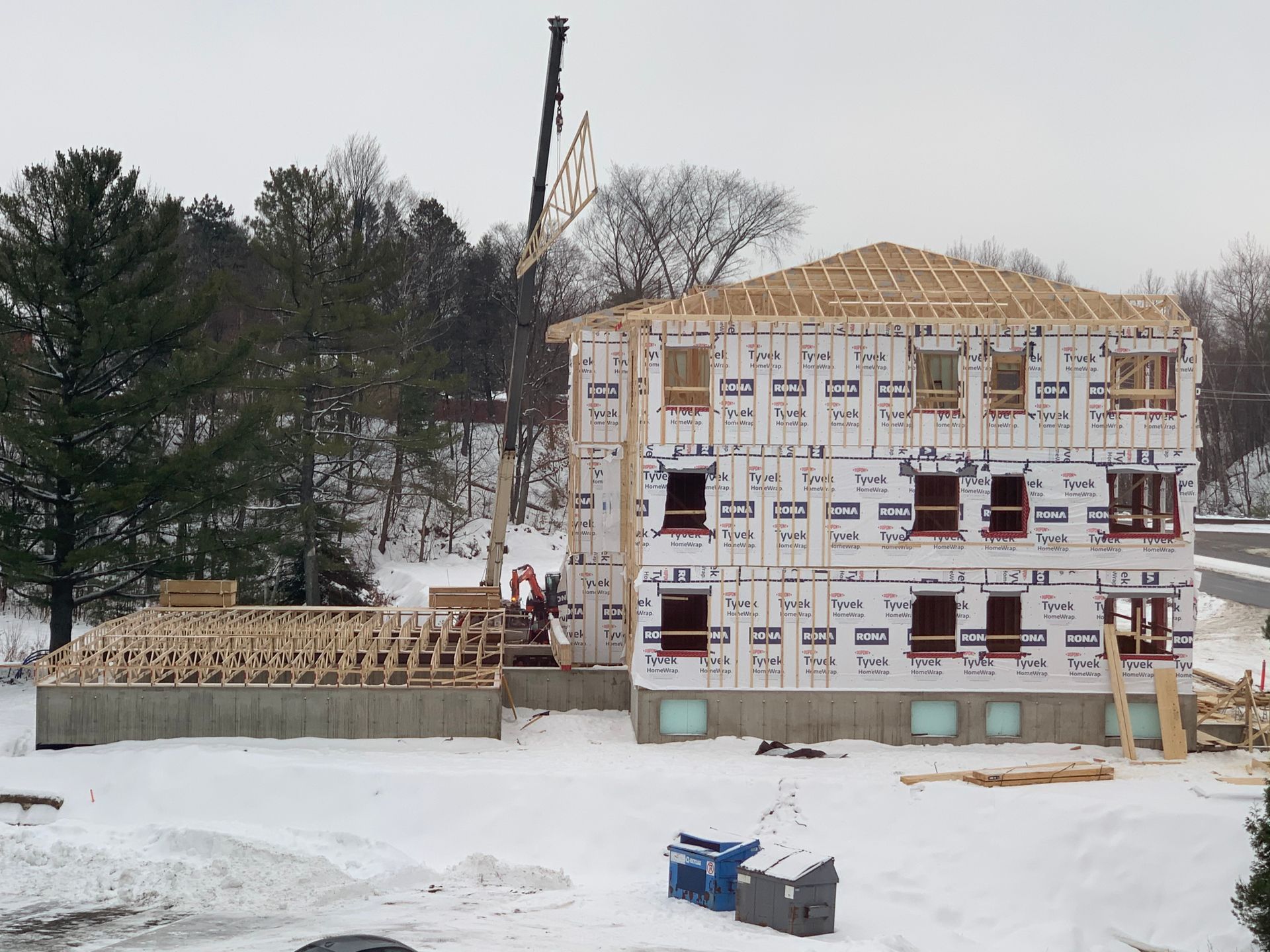 Chantier de construction en hiver. Une grue soulève la charpente d'un bâtiment de trois étages en construction. Sol enneigé.