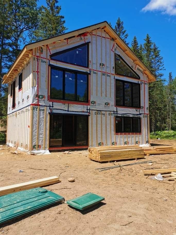 Maison de deux étages en construction, avec de grandes fenêtres. Les murs extérieurs ne sont pas finis, dans une zone boisée.
