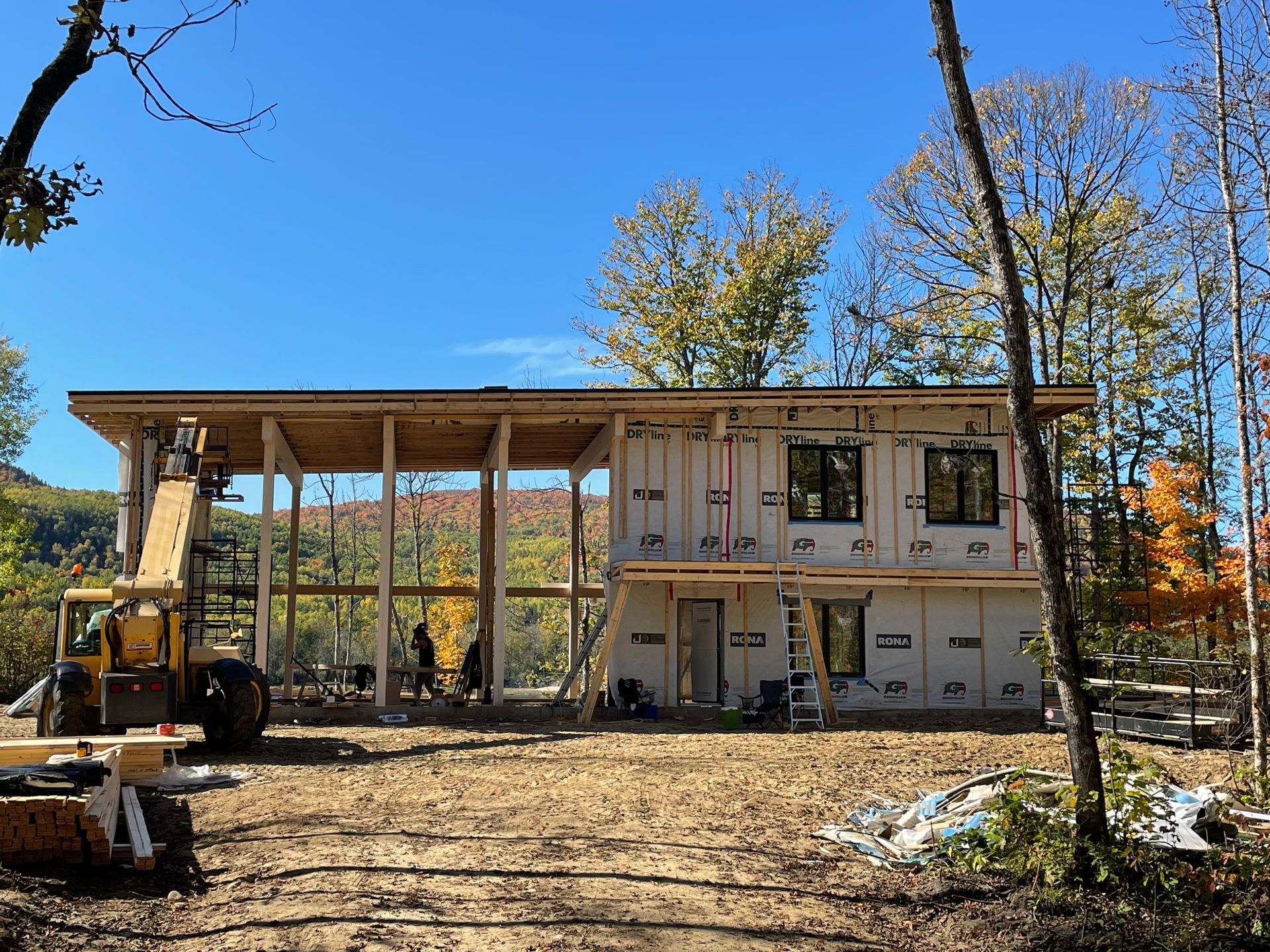 Construction d'une maison en bois à deux étages sous un ciel bleu, avec des ouvriers, une grue et des arbres environnants.