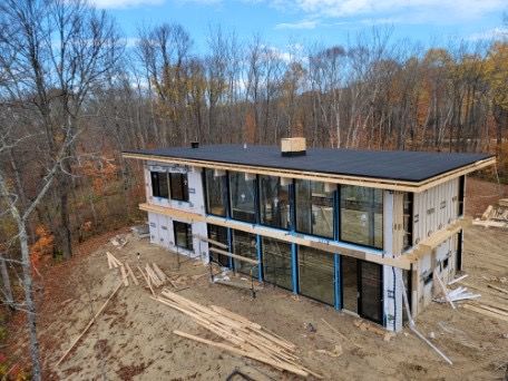 Maison moderne de deux étages en construction ; grandes fenêtres, toit noir, bardage en bois et arbres environnants.