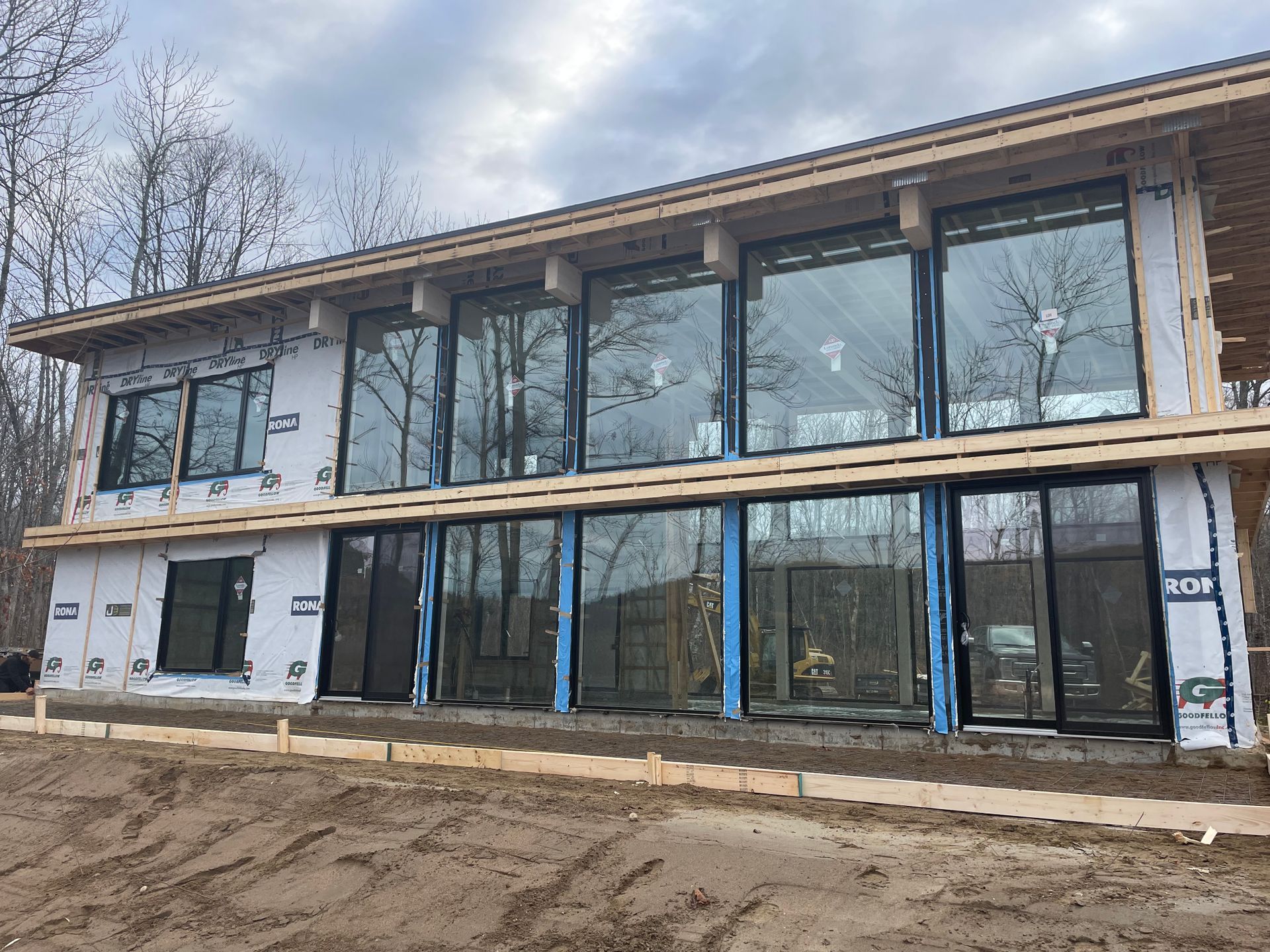 Maison à deux étages en construction, avec de grandes fenêtres. Murs extérieurs recouverts d'un film bleu. Terre brune et arbres en arrière-plan.