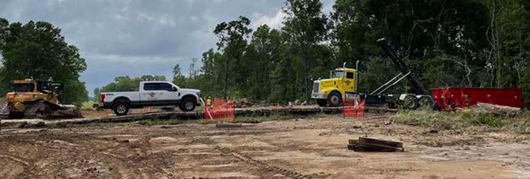Construction vehicles on a muddy site, including a white pickup, yellow excavator, and red dump truck.