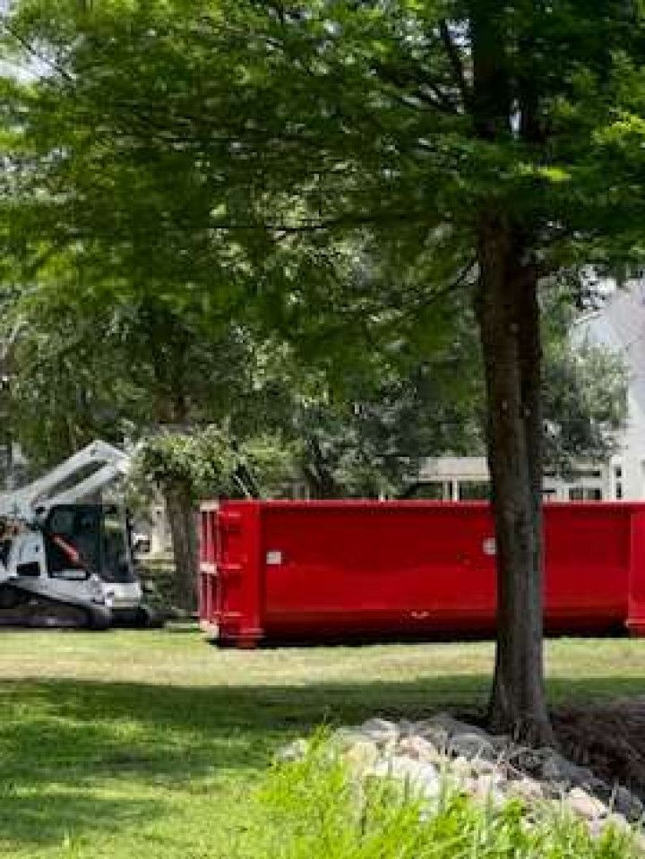 Red dumpster beside a white excavator in a grassy yard under leafy trees.