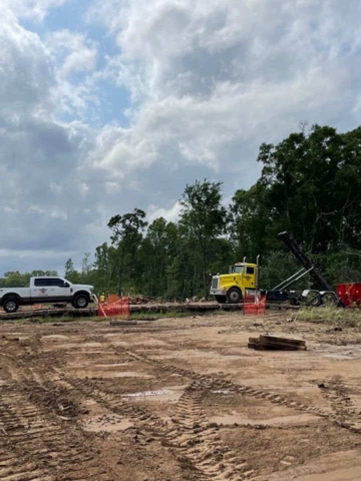 Construction site with muddy ground, white pickup truck, yellow excavator, and red barriers under a cloudy sky