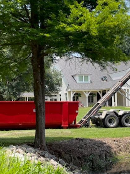 Red construction dumpster beside a house with a truck, partly obscured by a tree.