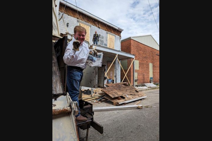 Person standing on debris in a narrow alley beside damaged buildings, holding papers.