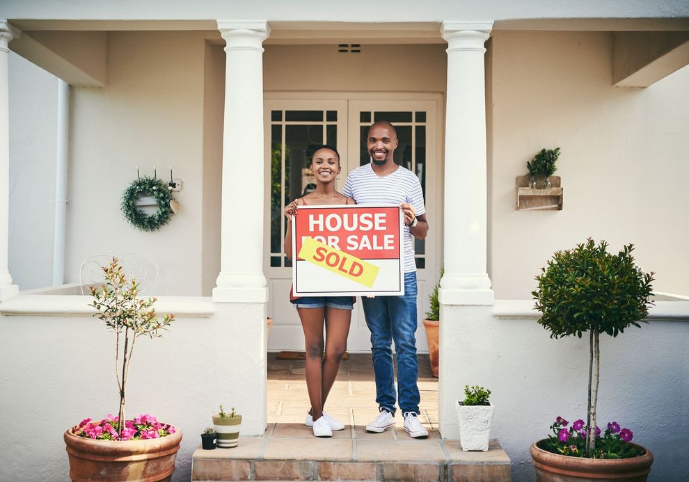 Couple standing at a front door holding a “House Sold” sign on a house porch.
