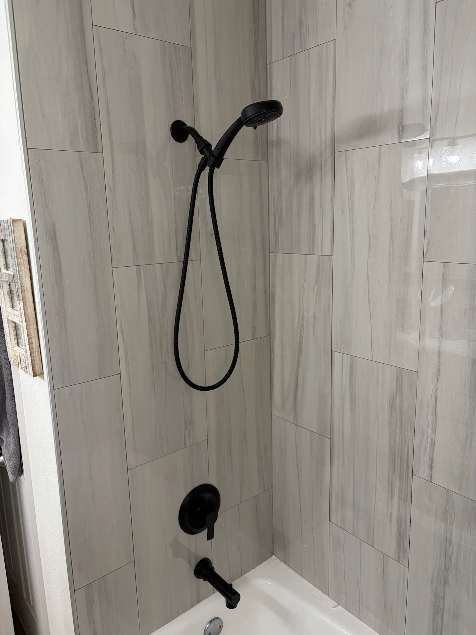 Black shower fixture with handheld wand in a tiled bathroom shower stall.