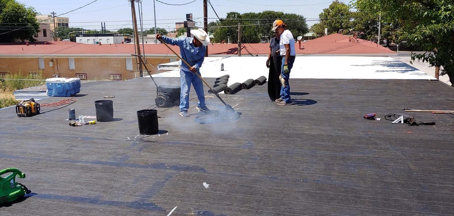 Workers spraying a dark rooftop coating on a flat roof with buckets and tools nearby