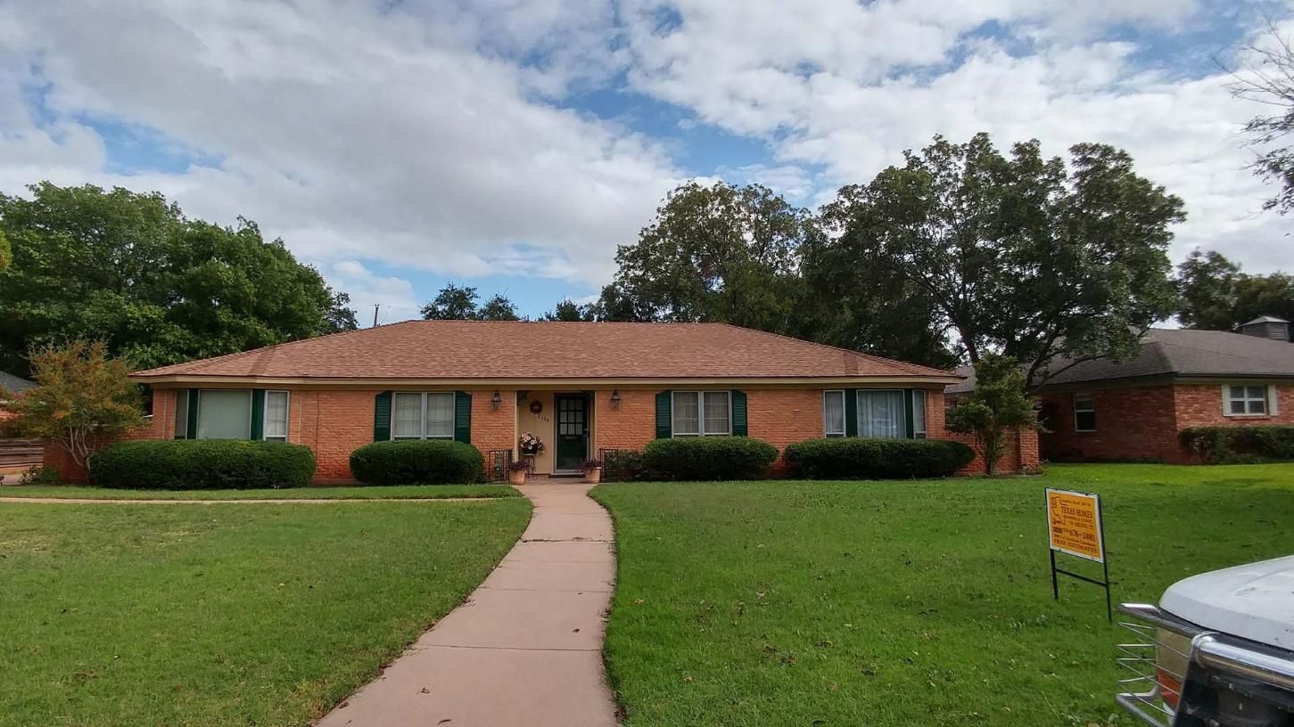 Single-story brick house with green lawn, walkway, and trees under a cloudy sky