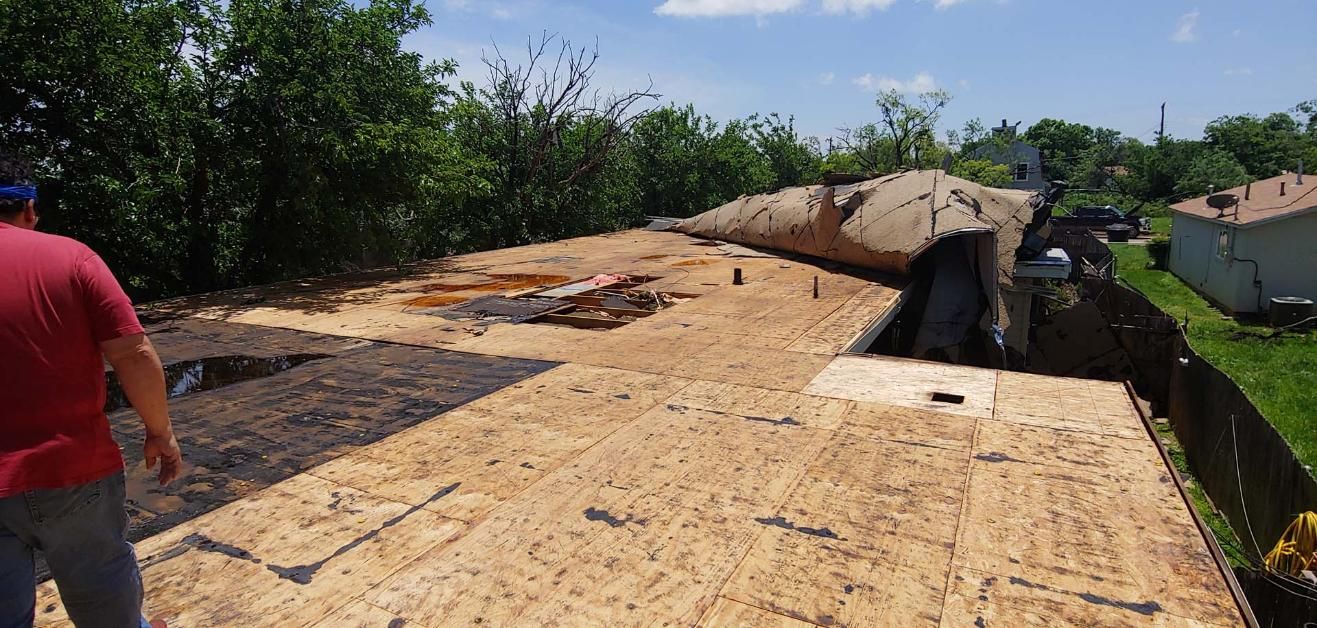 Damaged roof with tarps and debris beside green yard and houses under a bright sky