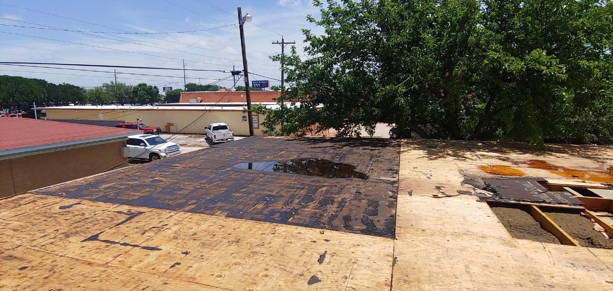 Rooftop with peeled-up roofing material and debris, next to trees and parked cars under a bright sky