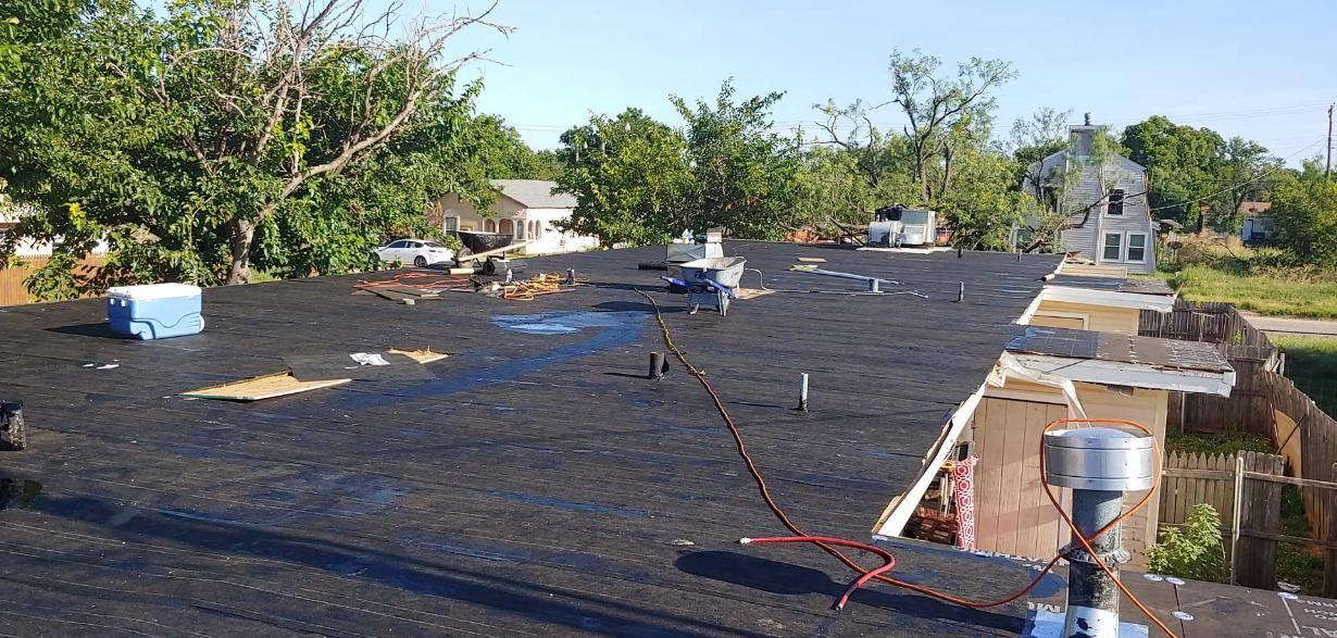 Rooftop covered in black roofing material with scattered tools and buckets, surrounded by trees and nearby houses.