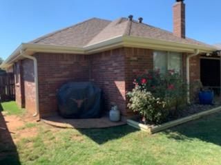 Brick house with a covered patio, brick chimney, and a grassy yard with shrubs.