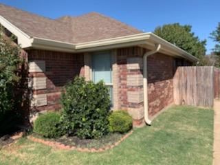 Brick house corner with front shrubs, a small lawn, and a wooden privacy fence under a blue sky