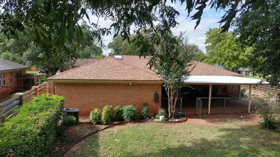 Backyard view of a brick house with a covered patio, trees overhead, and a grassy yard