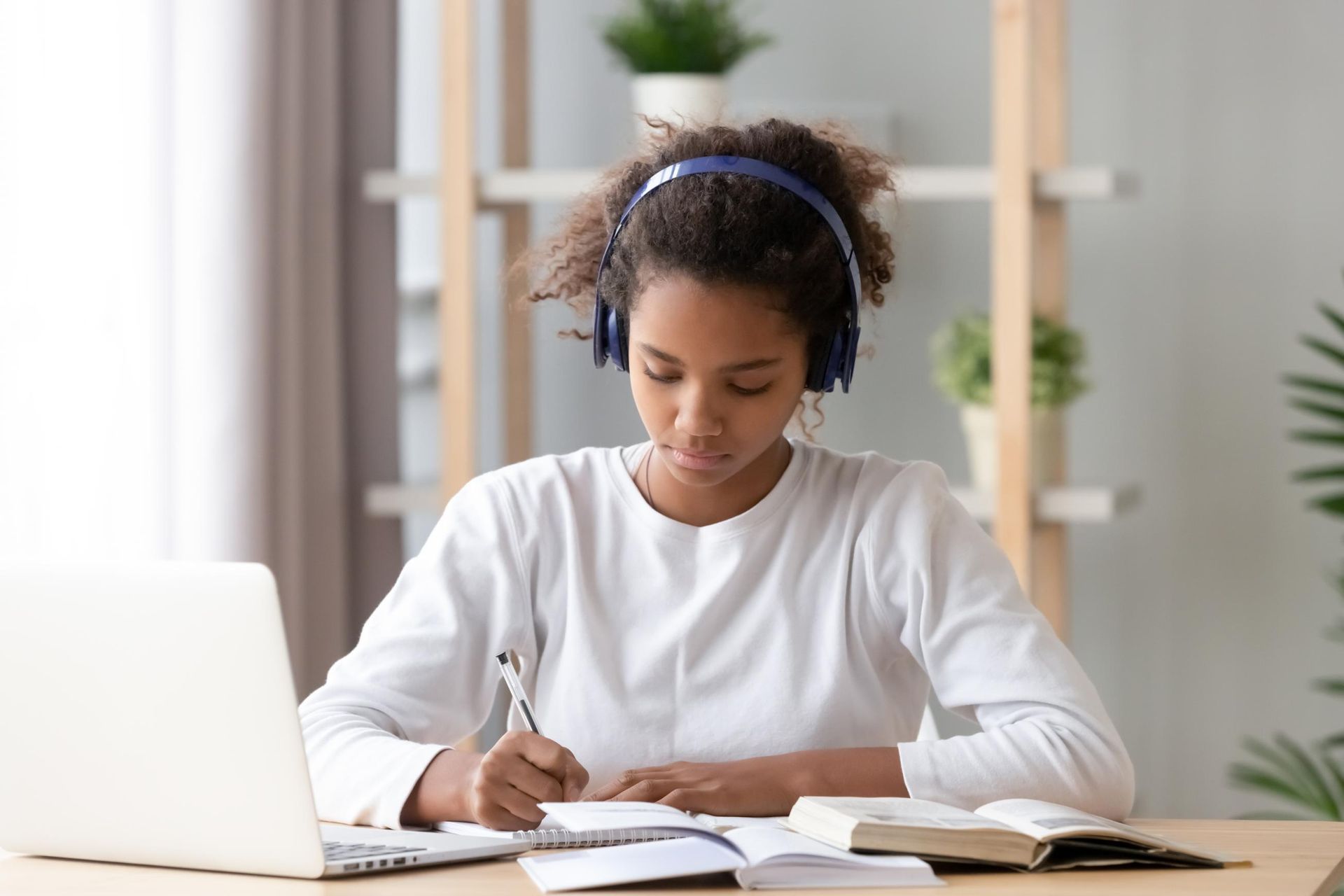 Person writing at a desk beside an open laptop and books in a bright home workspace