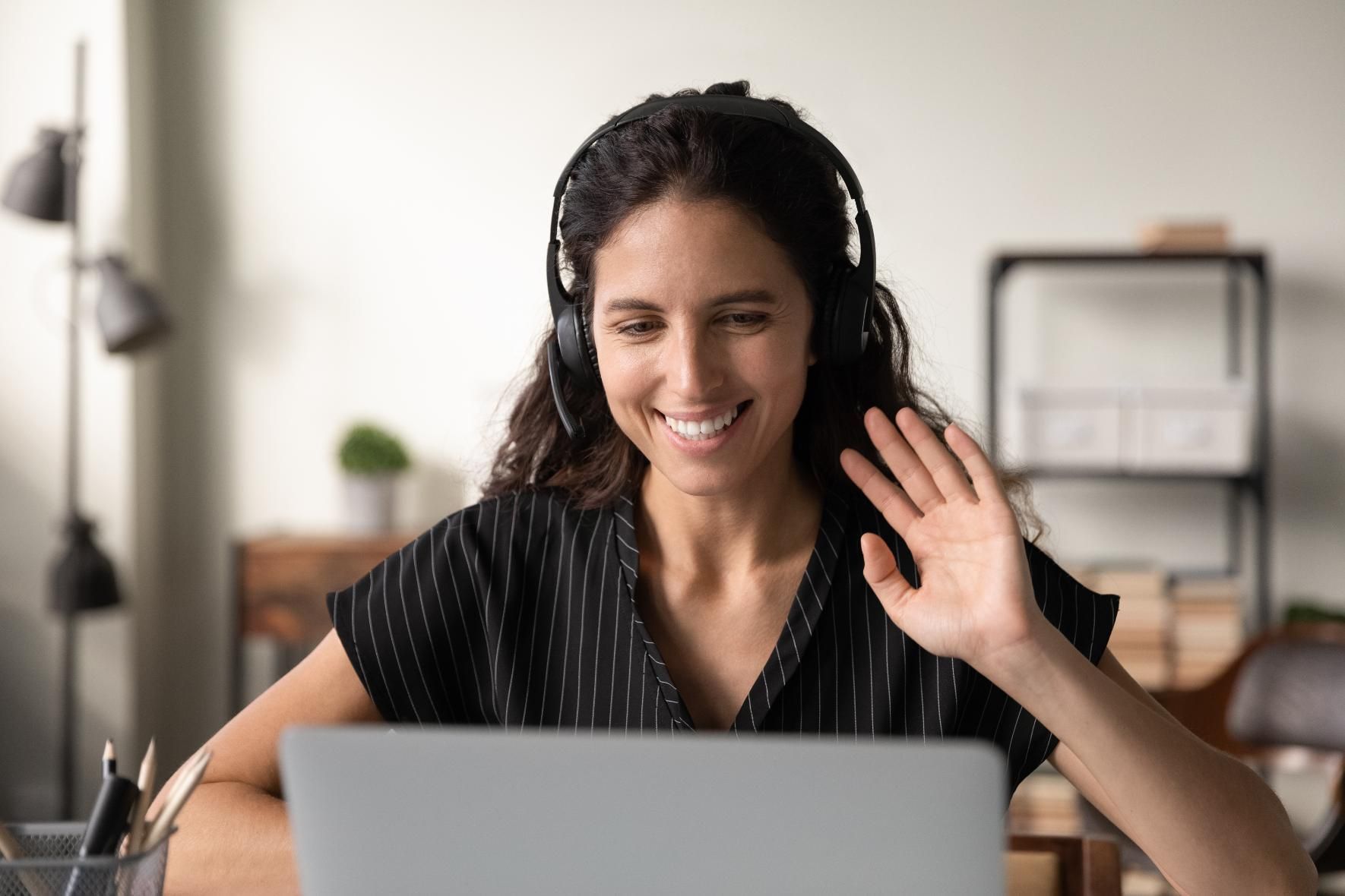 Woman smiling and waving at a laptop during a video call in a bright home office.