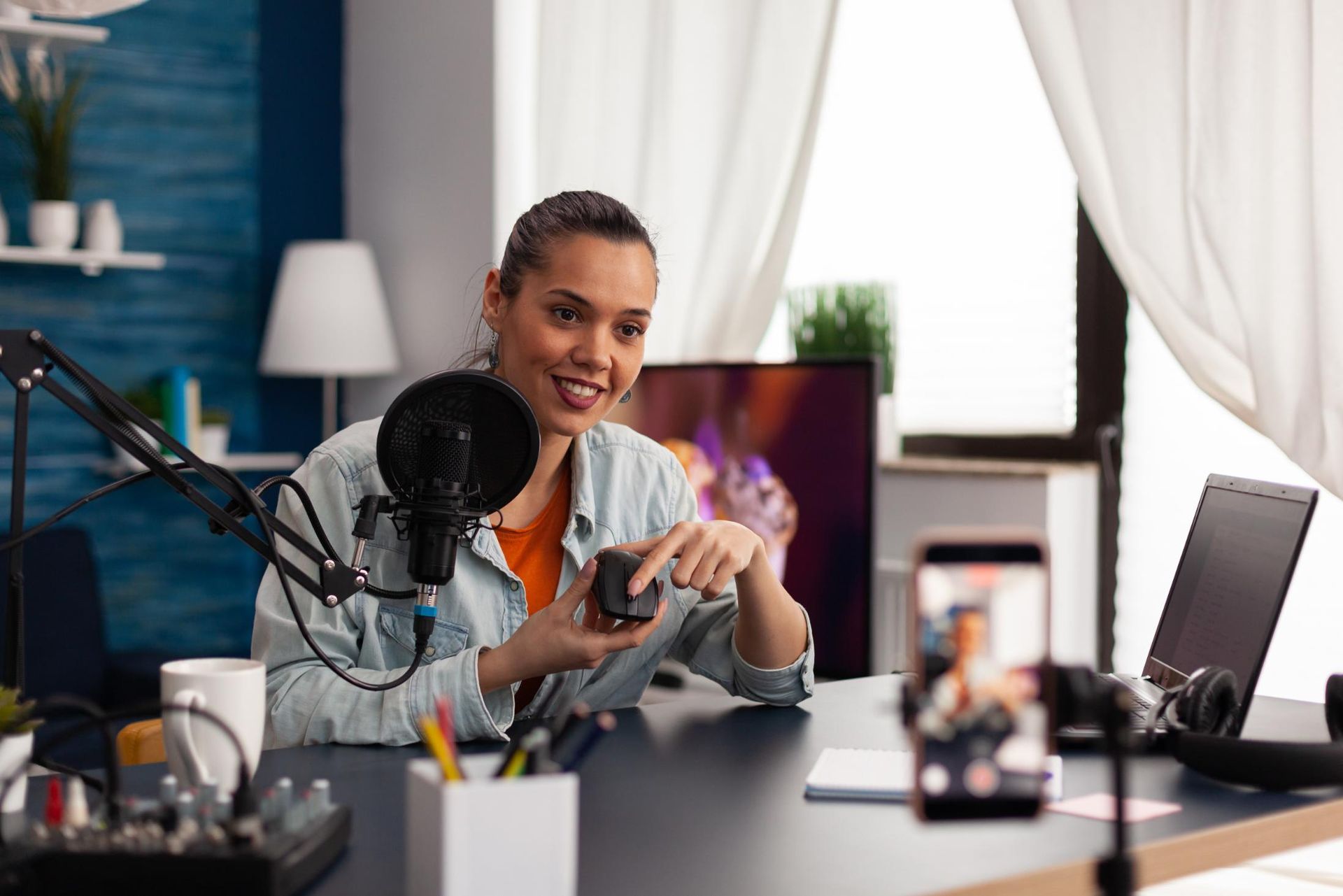 Person speaking into a microphone at a desk with a laptop, in a bright home studio