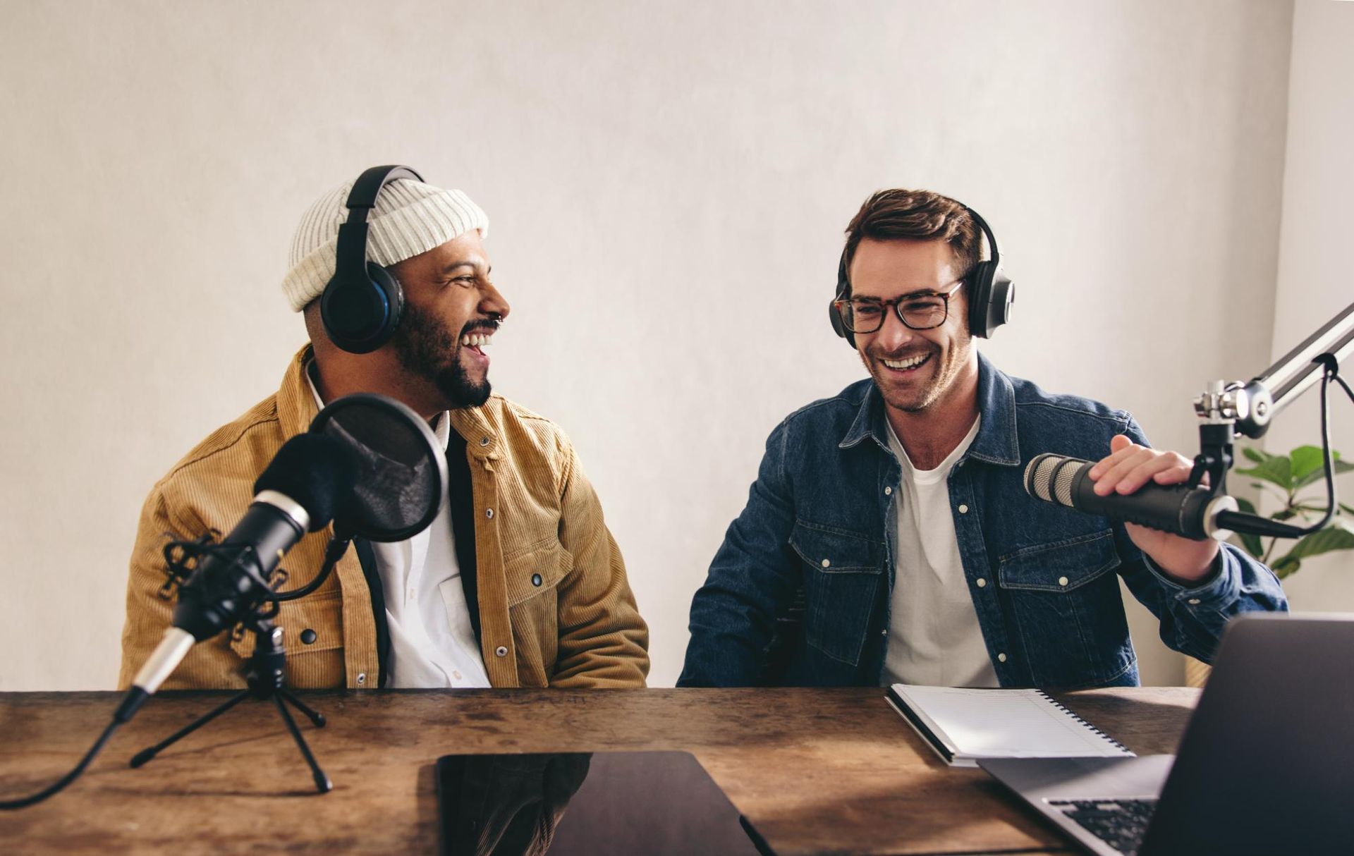 Two people recording a podcast at a desk with microphones, a laptop, and headphones