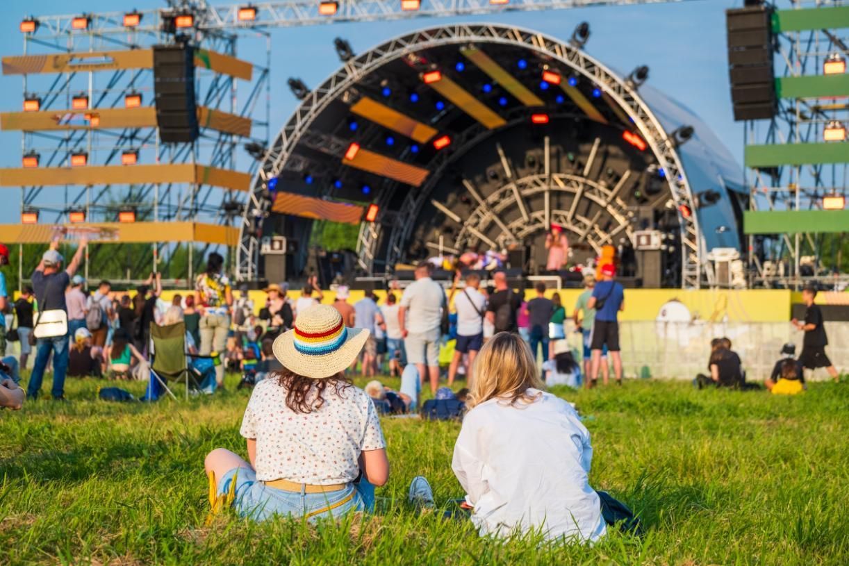 People sitting on grass facing a large outdoor concert stage at a festival