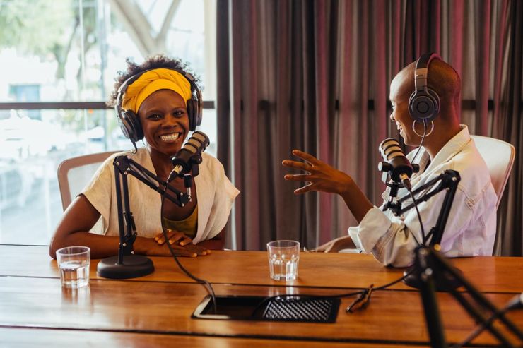 Two people recording a podcast at microphones in a studio, smiling across a table.