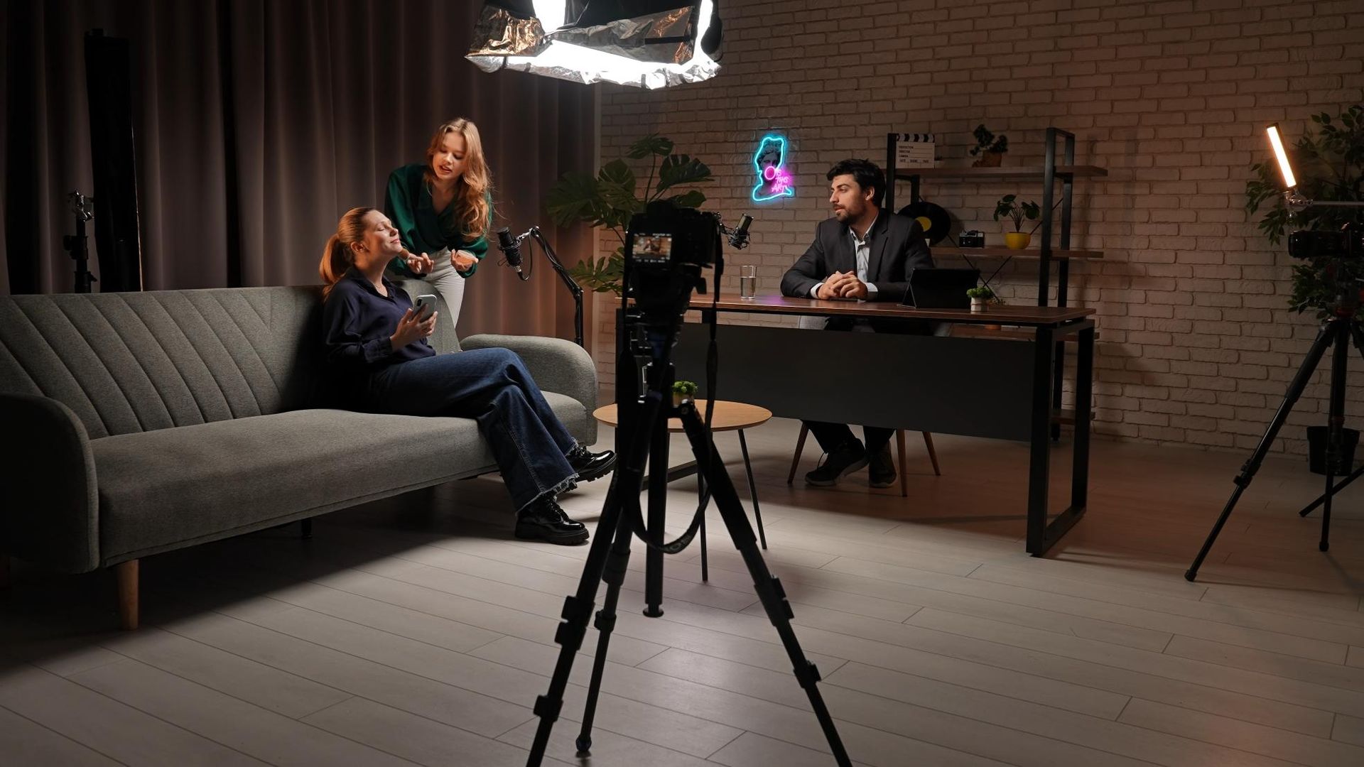 Podcast studio with two people on a couch and a host at a desk, lit by studio lights and cameras