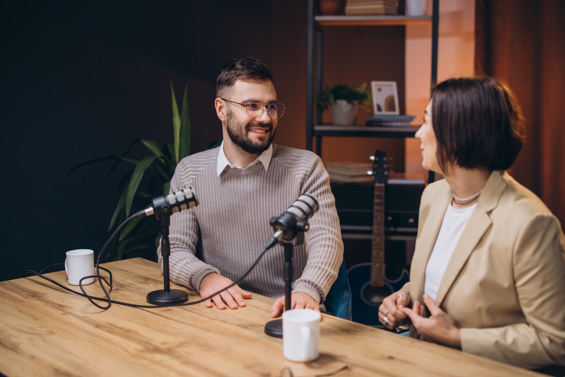 Two people seated at microphones in a podcast studio, smiling and talking across a wooden table.