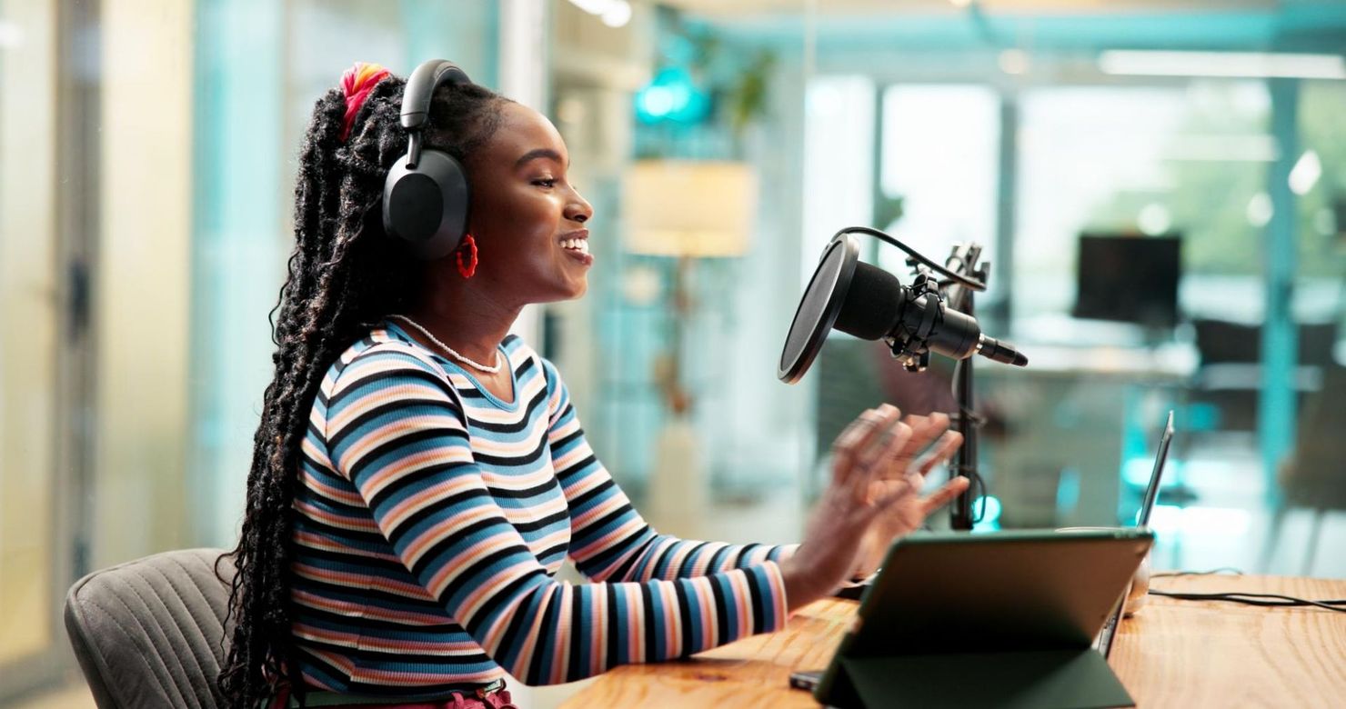 Podcast host wearing headphones speaks into a studio microphone at a desk with a laptop.