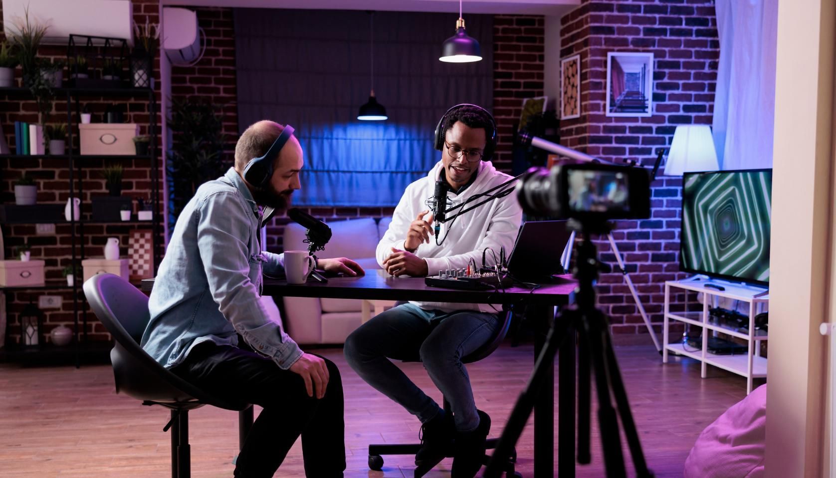 Two people in a studio discussing at a table, with a camera filming them under purple lighting.
