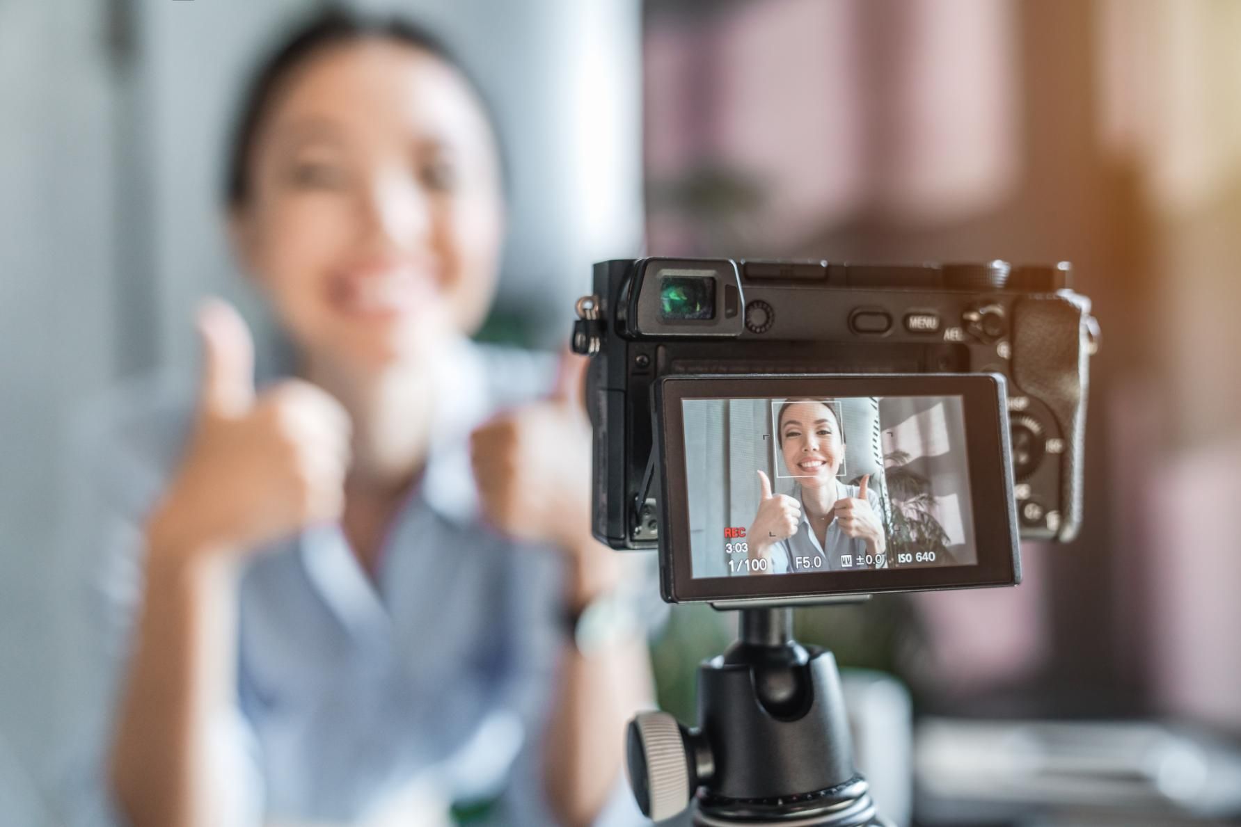 Camera screen showing a smiling person giving a thumbs-up on a tripod in a blurred indoor setting