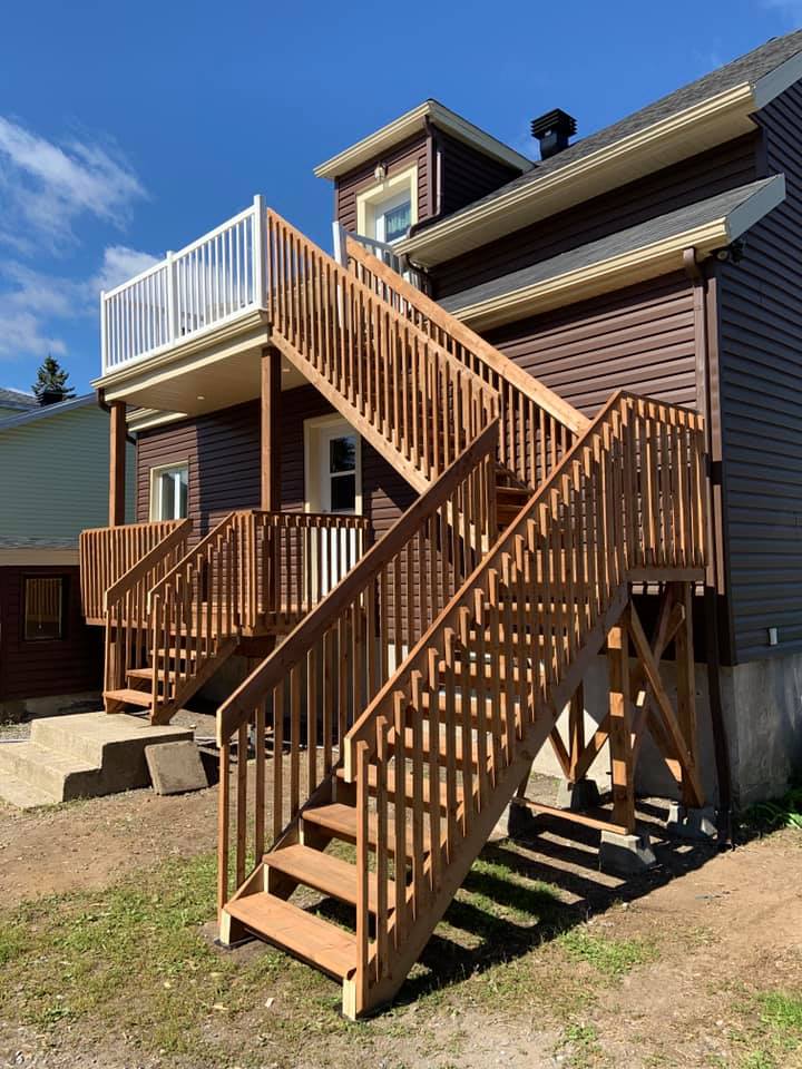 Un escalier extérieur en bois mène à un bâtiment brun avec un balcon blanc, par une journée ensoleillée.