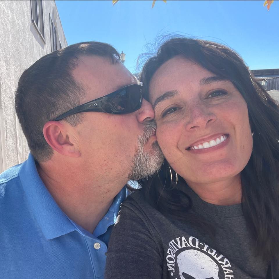 Man in sunglasses kisses smiling woman outdoors under a bright blue sky.