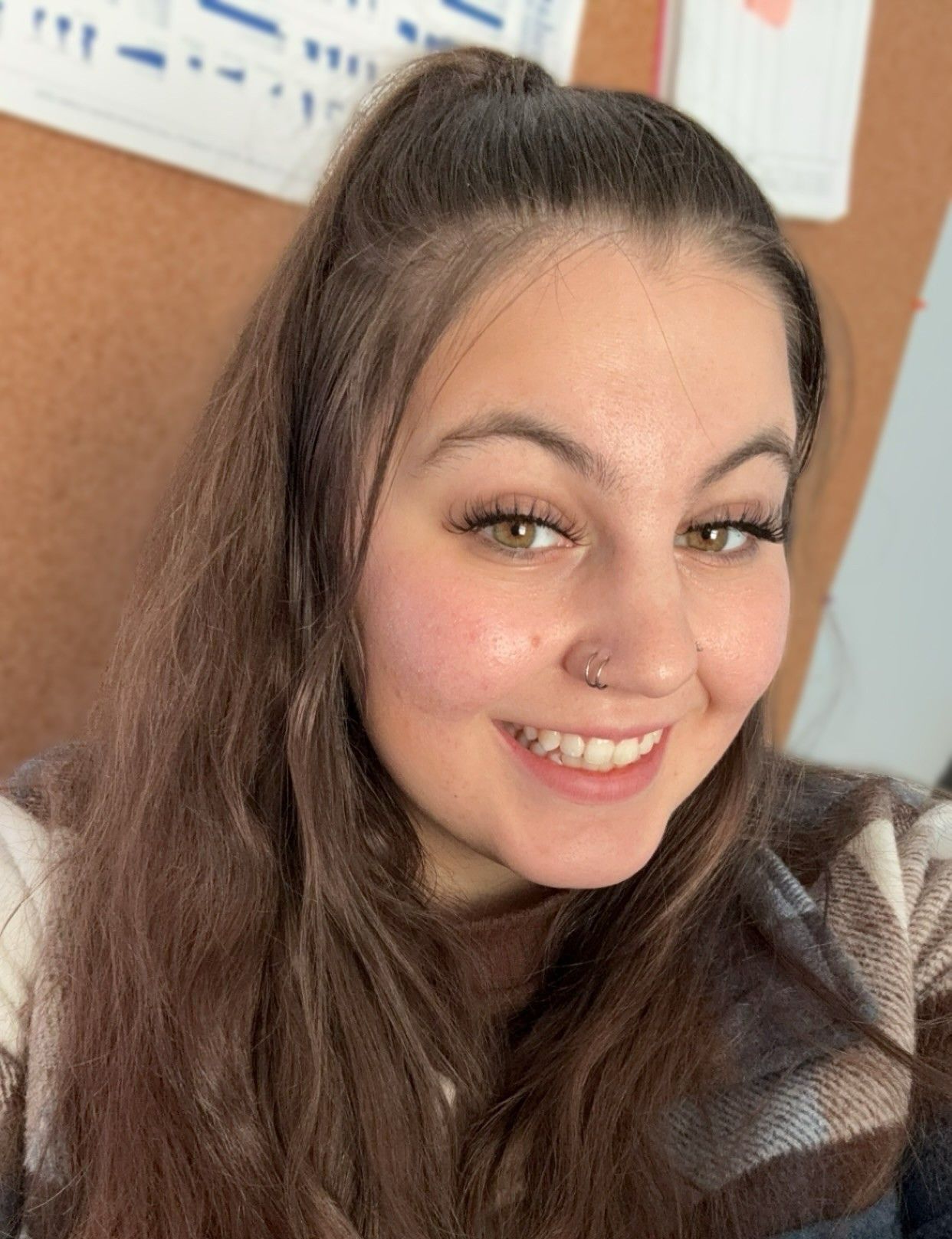 Smiling person with long brown hair indoors, wearing a plaid shirt against a light-colored wall.